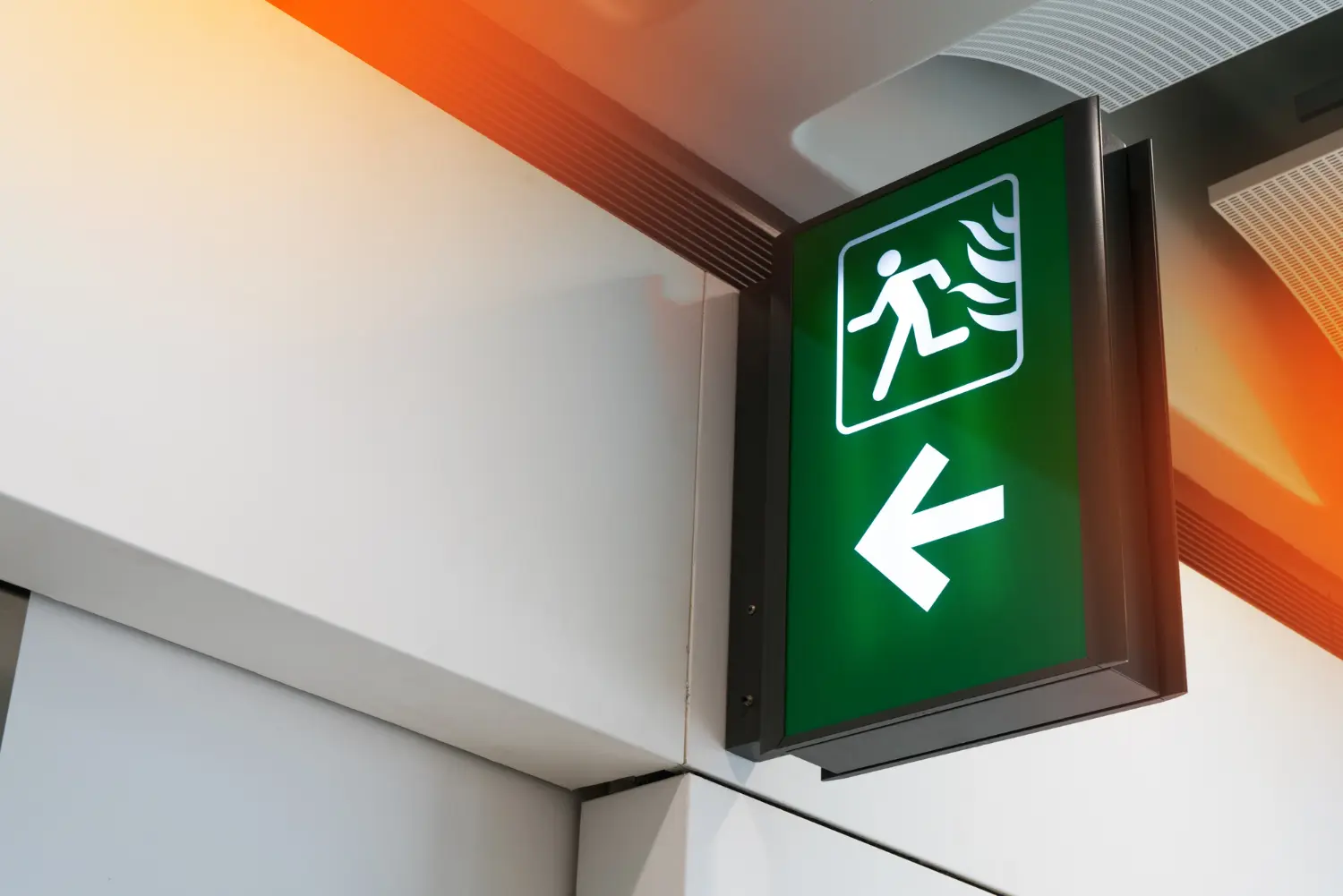 Placa suspensa de emergência iluminada com seta direcional esquerda, instalada em nível superior e ângulo de visada lateral. Elemento essencial para evacuação eficiente conforme NBR 16820, especialmente em áreas amplas e corredores.