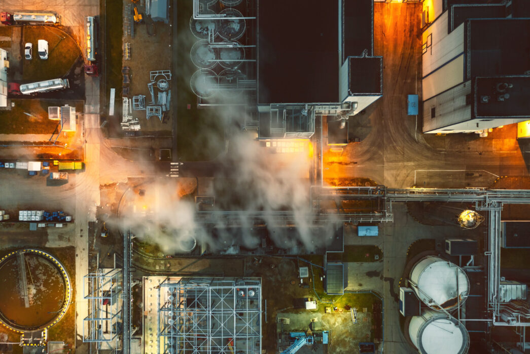 Vista aérea de uma instalação industrial fervilhante, com vapores escapando das tubulações e uma iluminação dramática, criando uma sensação de instabilidade em um ambiente potencialmente explosivo.