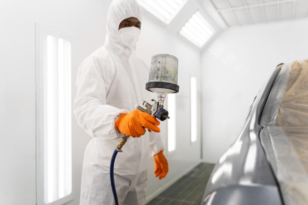 Aplicação de tinta em ambiente controlado com cabine pressurizada. Operador devidamente paramentado com EPI completo (macacão, máscara, luvas). Iluminação uniforme e paredes limpas indicam conformidade com boas práticas de pintura automotiva. - Laudo Cabine Pintura.