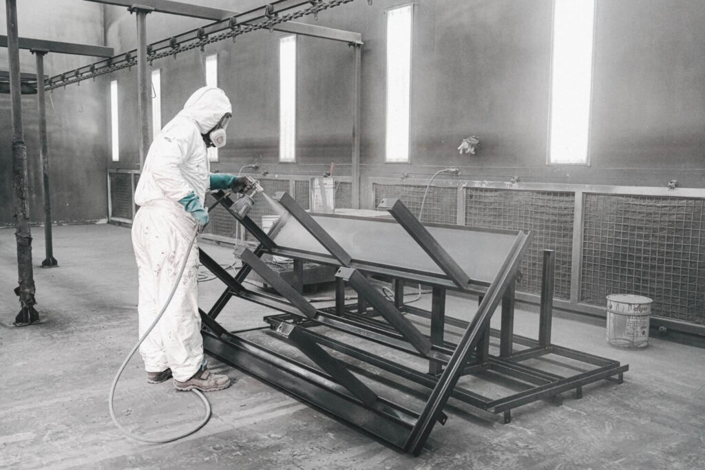 Pintura industrial de estruturas metálicas em cabine com exaustão e ventilação forçada. Operador com máscara facial adequada, demonstrando controle de exposição a névoas de tinta. Piso apresenta vestígios de acúmulo de tinta, recomendando-se limpeza periódica conforme norma. - Laudo Cabine Pintura.