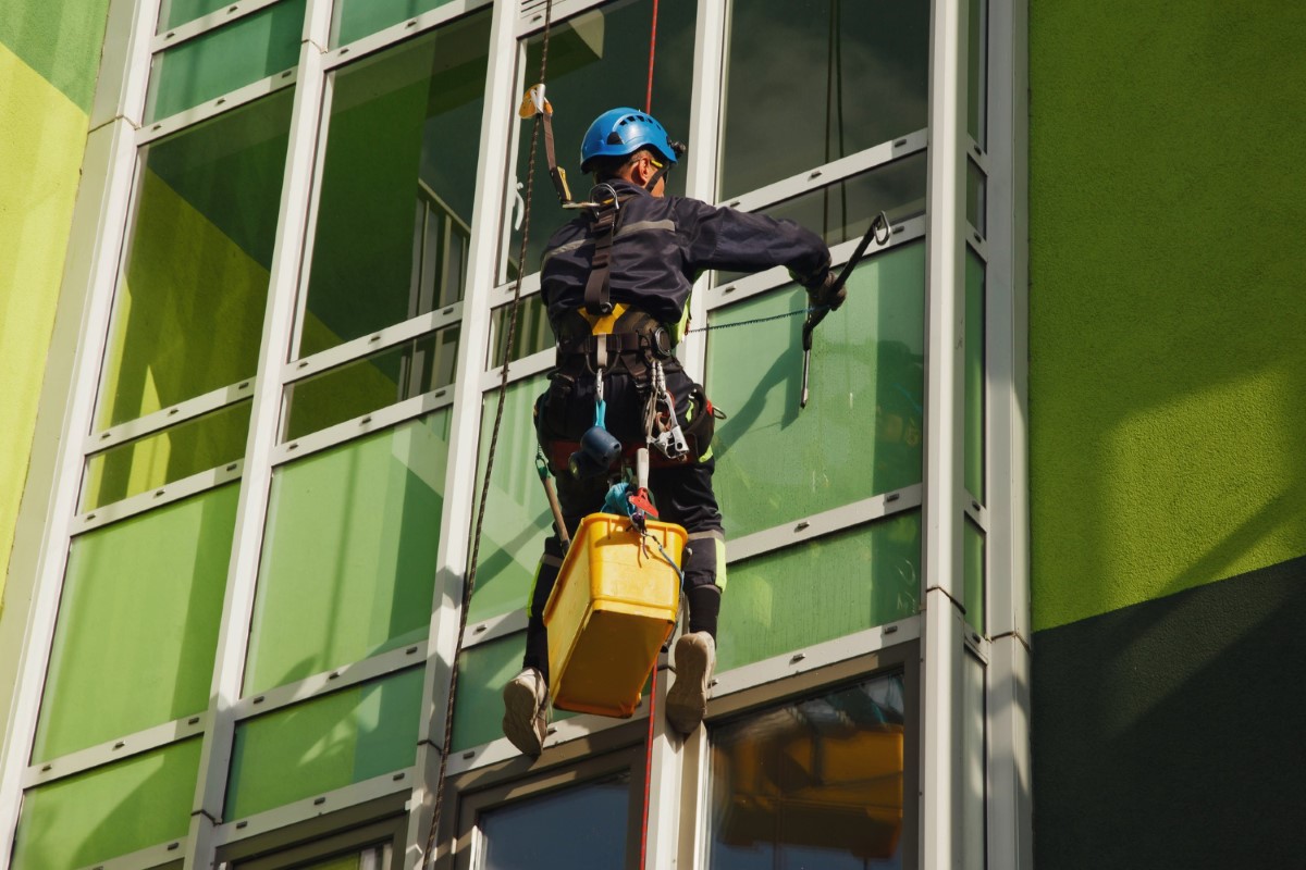Worker operates a bosun chair while performing façade maintenance, maintaining full control of descent and anchorage stability according to NR 18 requirements.