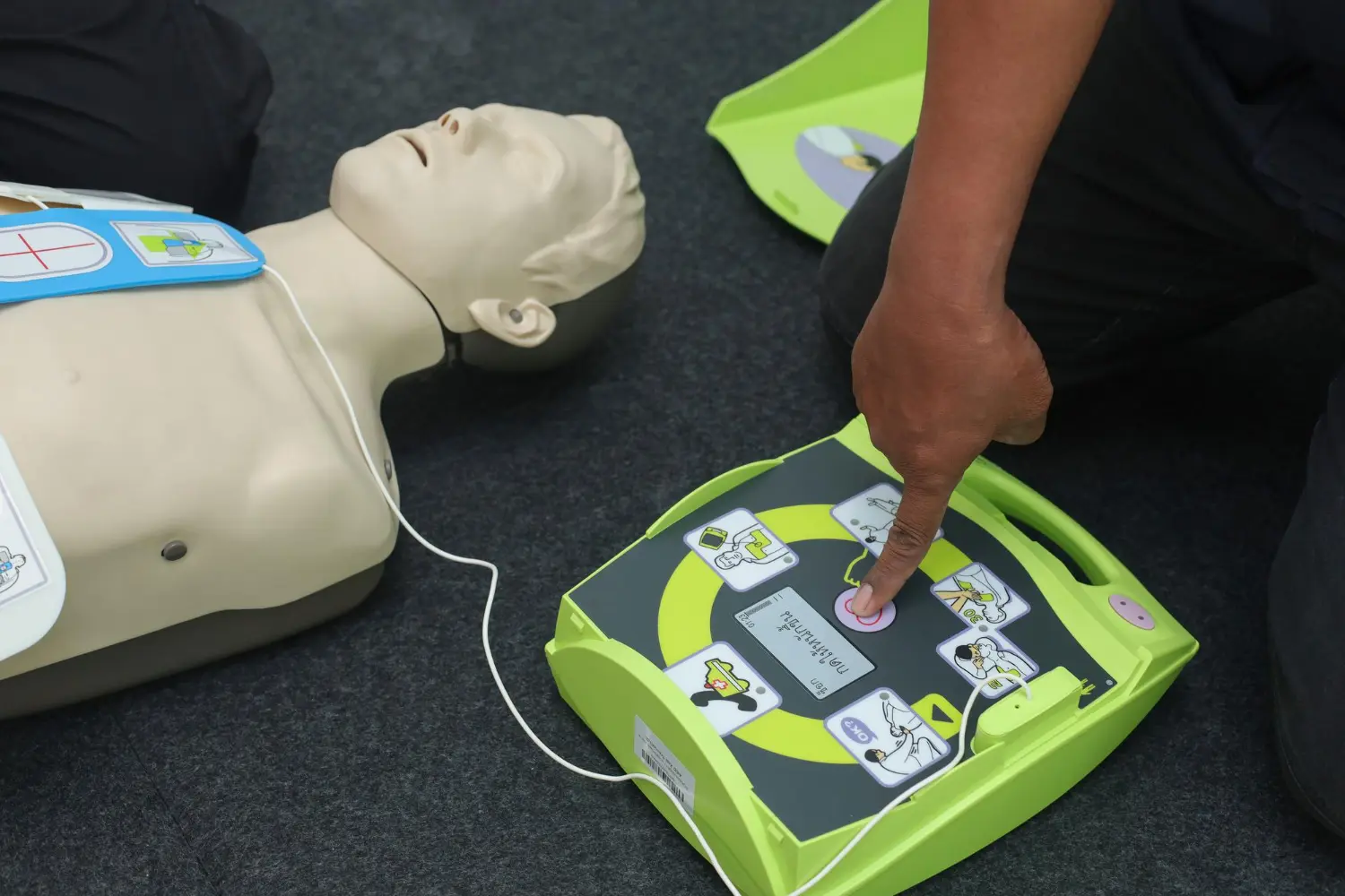 Simulação de atendimento com DEA em adulto operador aciona o botão de choque após análise automática do ritmo cardíaco. Equipamento posicionado corretamente ao lado da vítima.