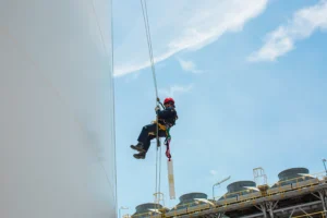 Profissional executa atividade em altura utilizando técnicas de acesso por linha de vida, mantendo posicionamento seguro para inspeção e manutenção em superfície vertical de tanque industrial.