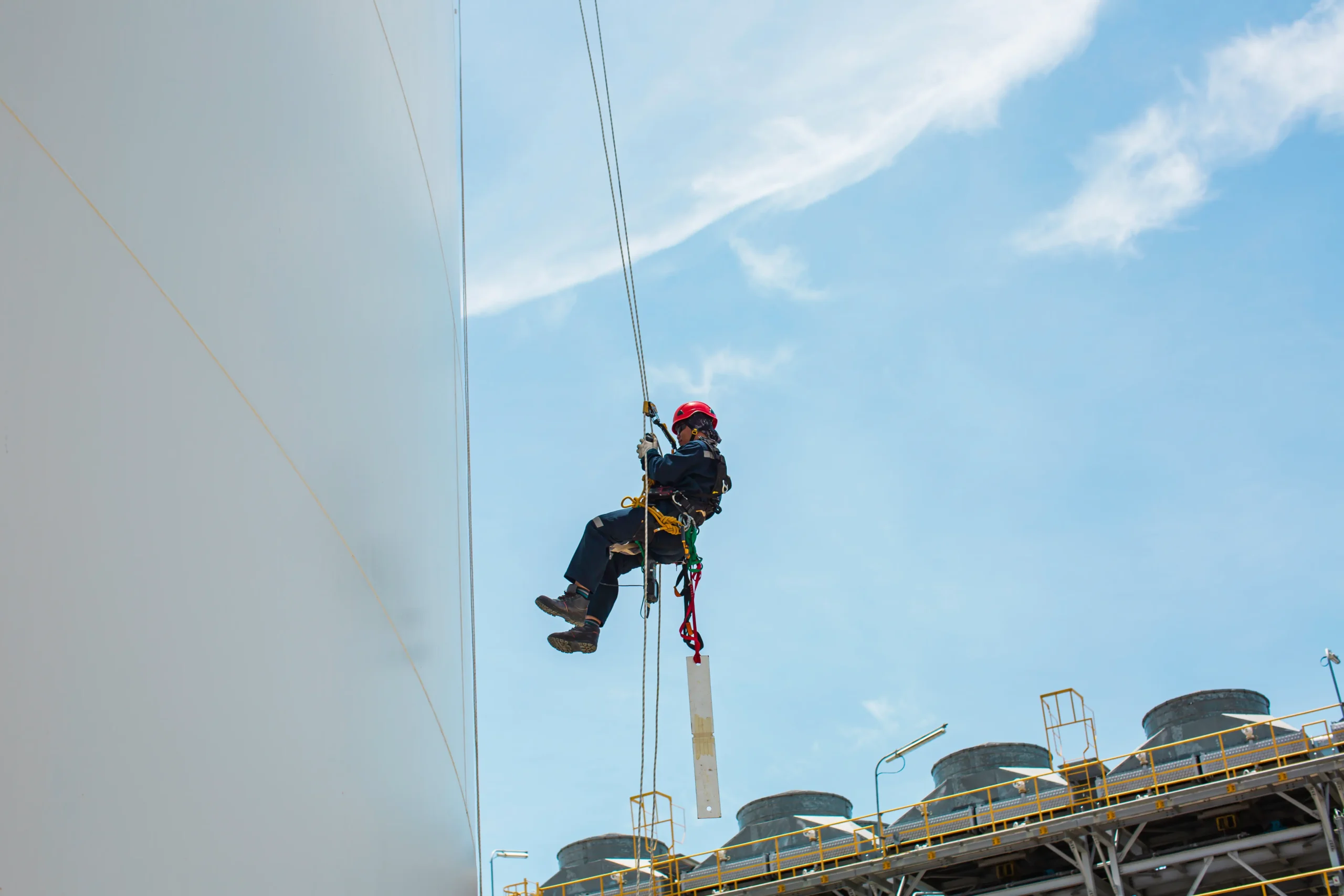 Profissional executa atividade em altura utilizando técnicas de acesso por linha de vida, mantendo posicionamento seguro para inspeção e manutenção em superfície vertical de tanque industrial.