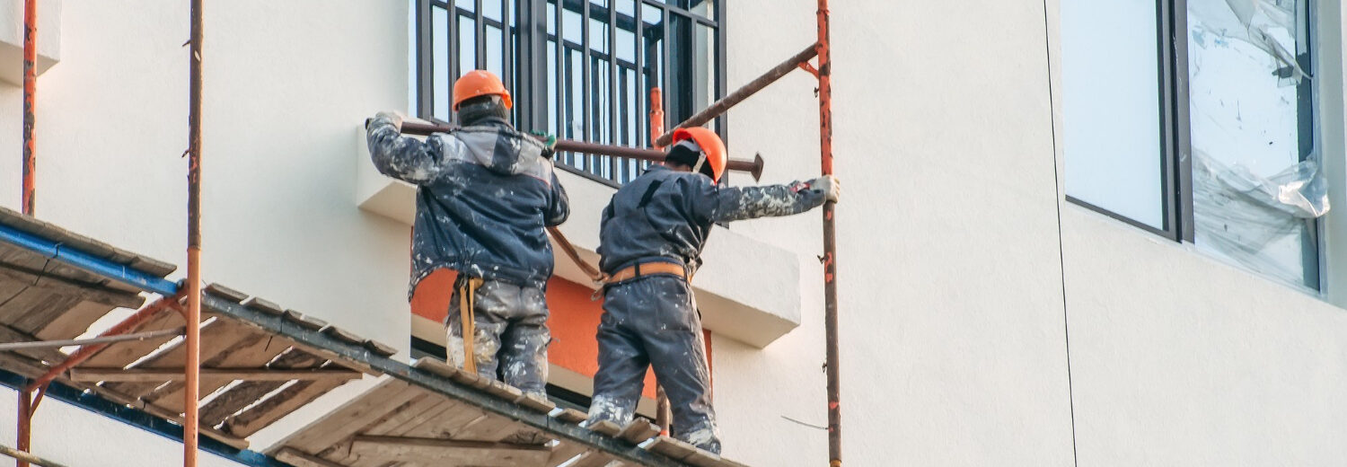 Trabalhadores realizam montagem de andaime em fachada de edifício, atuando em altura com uso de capacetes e vestimentas de proteção - Curso Acesso por Corda 2 NBR 15475