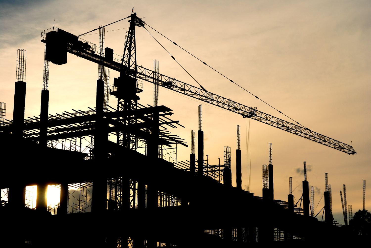 Estrutura de um edifício em construção ao entardecer, com guindastes e armações de aço destacando-se no horizonte, representando o progresso e a engenharia moderna. Curso NR 18 Inicial.