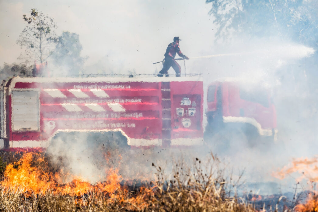 Um bombeiro em cima de um caminhão de combate a incêndios usa um jato de água para conter chamas intensas em uma área de vegetação. Isso destaca a importância da resposta rápida e uso de equipamentos adequados para prevenir a propagação de incêndios em instalações agrícolas. - Curso NFPA 61.