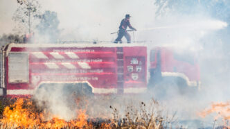 Um bombeiro em cima de um caminhão de combate a incêndios usa um jato de água para conter chamas intensas em uma área de vegetação. Isso destaca a importância da resposta rápida e uso de equipamentos adequados para prevenir a propagação de incêndios em instalações agrícolas. - Curso NFPA 61.