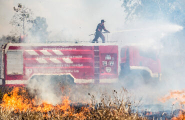 Um bombeiro em cima de um caminhão de combate a incêndios usa um jato de água para conter chamas intensas em uma área de vegetação. Isso destaca a importância da resposta rápida e uso de equipamentos adequados para prevenir a propagação de incêndios em instalações agrícolas. Um bombeiro em cima de um caminhão de combate a incêndios usa um jato de água para conter chamas intensas em uma área de vegetação. Isso destaca a importância da resposta rápida e uso de equipamentos adequados para prevenir a propagação de incêndios em instalações agrícolas. - Curso NFPA 61.