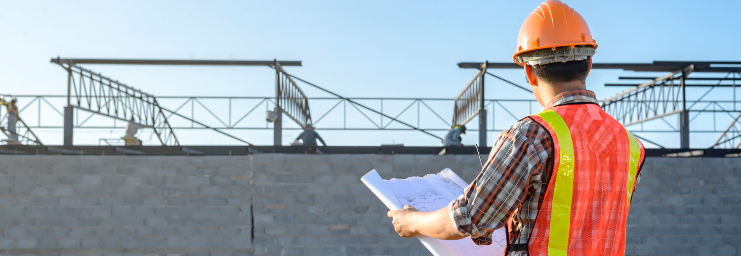 Um engenheiro ou supervisor de obra analisa plantas arquitetônicas em frente a um edifício em construção, assegurando que todos os detalhes estejam alinhados com o planejamento. - Curso Capacitação Incial NR 18. Curso NR 18 Inicial.