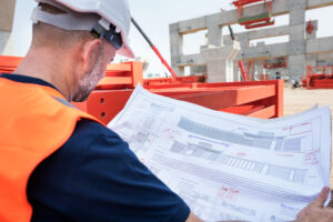 Engenheiro civil analisando o detalhamento executivo de uma estrutura de concreto armado, revisando seções de armaduras, especificações de fôrmas, e cotas do projeto no canteiro de obras. Essa etapa é crucial para verificar a compatibilidade entre o projeto estrutural e as condições reais de execução.