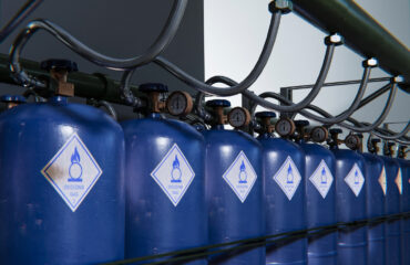 Cilindros de gás oxidante armazenados em série, interligados por tubulações de alta pressão e válvulas reguladoras. Os rótulos indicam a natureza do conteúdo, com potencial para p Cilindros de gás oxidante armazenados em série, interligados por tubulações de alta pressão e válvulas reguladoras. Os rótulos indicam a natureza do conteúdo, com potencial para promover combustão ao entrar em contato com materiais inflamáveis. Equipamentos assim são comuns em processos industriais e laboratoriais. - Curso Cilindro Gases Inflamáveis.