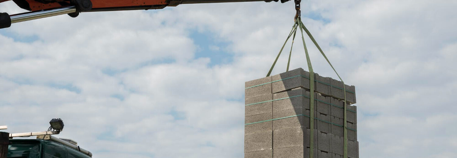 caminhão equipado com guindaste articulado realizando o içamento de um pallet contendo blocos de concreto
