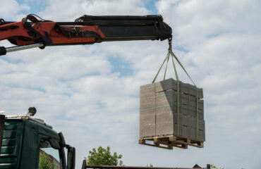 FOTO 01 caminhão equipado com guindaste articulado realizando o içamento de um pallet contendo blocos de concreto