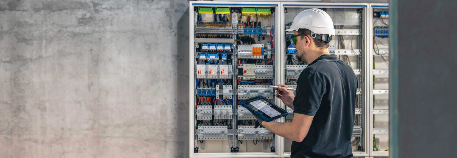 Técnico inspecionando um quadro de distribuição elétrica com auxílio de um tablet, assegurando a conformidade com o padrão de entrada de energia e a segurança na instalação. - Curso Padrão Entrada Energia.