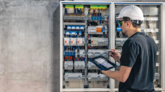 Técnico inspecionando um quadro de distribuição elétrica com auxílio de um tablet, assegurando a conformidade com o padrão de entrada de energia e a segurança na instalação. - Curso Padrão Entrada Energia.