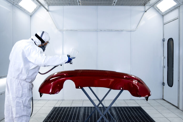 Inspeção final em peça automotiva recém-pintada. Ambiente iluminado e fechado, ideal para acabamento e controle visual de qualidade. Operador com EPI e em posição técnica de análise. Condições adequadas de luminosidade e limpeza observadas.