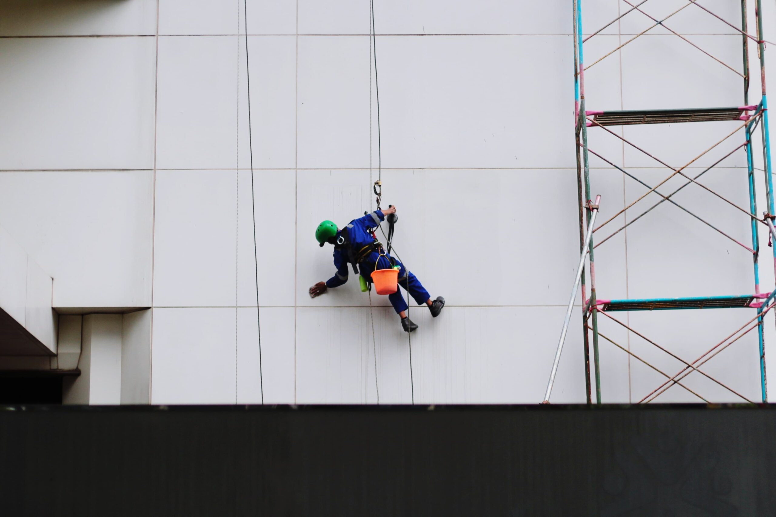 Profissional realiza atividade em fachada vertical com técnica de acesso por corda, utilizando capacete, talabarte, assento e ponto de ancoragem.