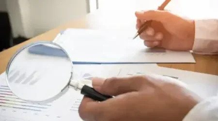 Profissional fazendo auditoria preventiva enquanto analisa dados de relatórios com uma lupa, calculadora e caneta.