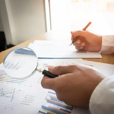 Profissional fazendo auditoria preventiva enquanto analisa dados de relatórios com uma lupa, calculadora e caneta.