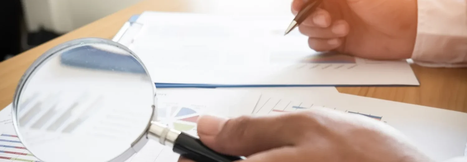 Profissional fazendo auditoria preventiva enquanto analisa dados de relatórios com uma lupa, calculadora e caneta.