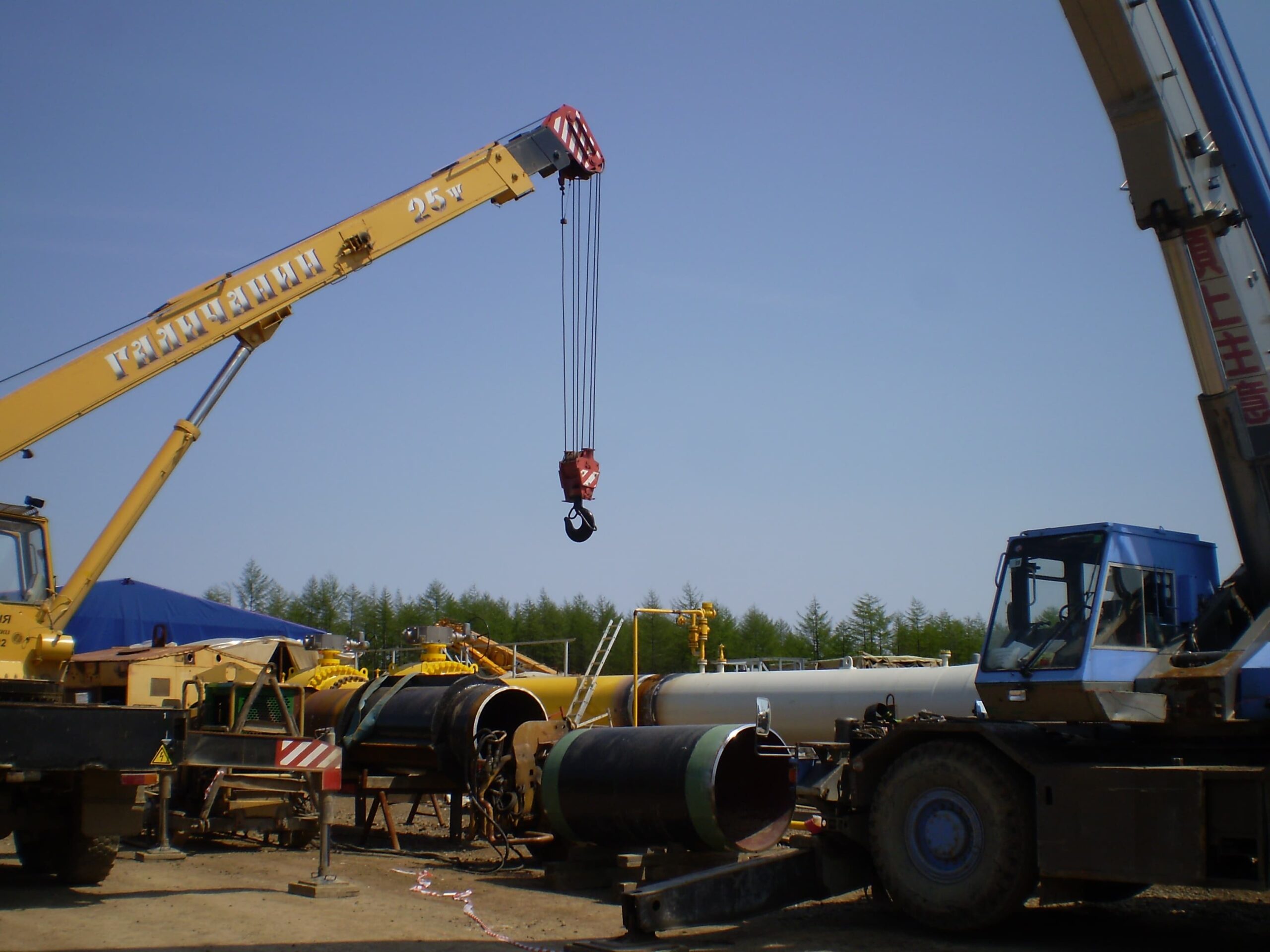 Dois guindastes hidráulicos de rodas realizam içamento de tubulações em área de construção de gasoduto. A operação exige controle absoluto do raio de alcance, análise prévia do solo e domínio técnico da tabela de carga.