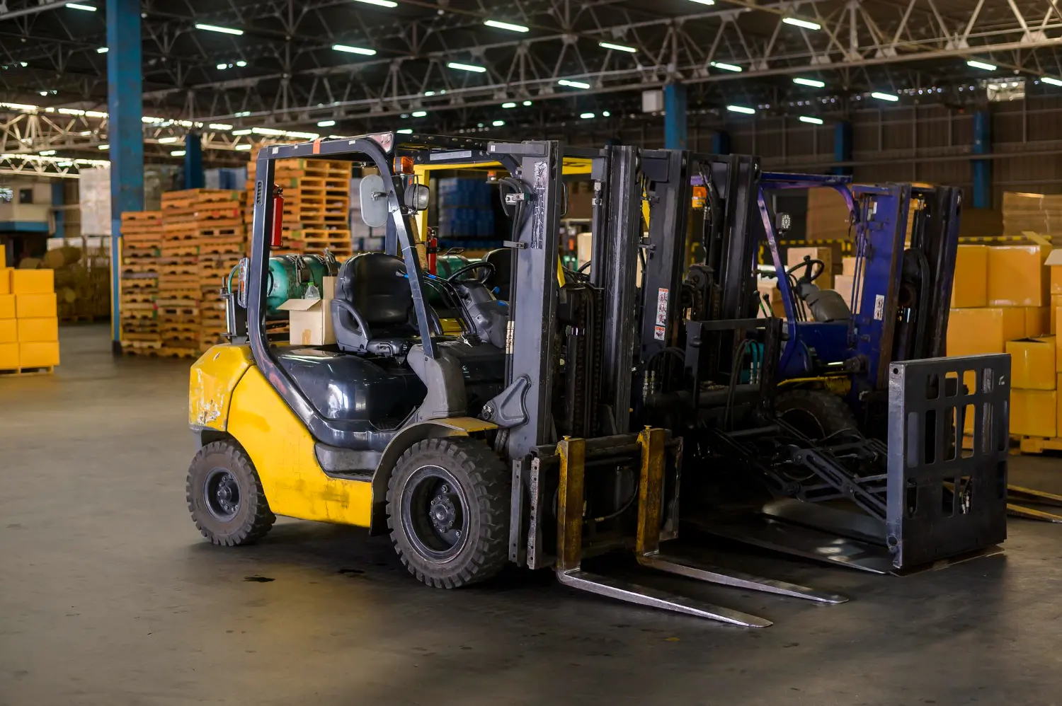 Empilhadeiras prontas para operação em área interna com pallets e equipamentos ao fundo.