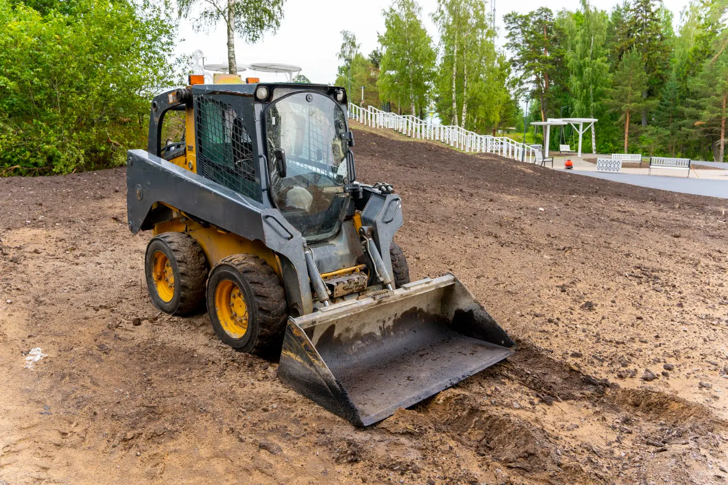 Mini carregadeira posicionada em aclive, pronta para escavação com controle de tração e estabilidade. A operação exige atenção aos riscos laterais e cumprimento das diretrizes da NR 11