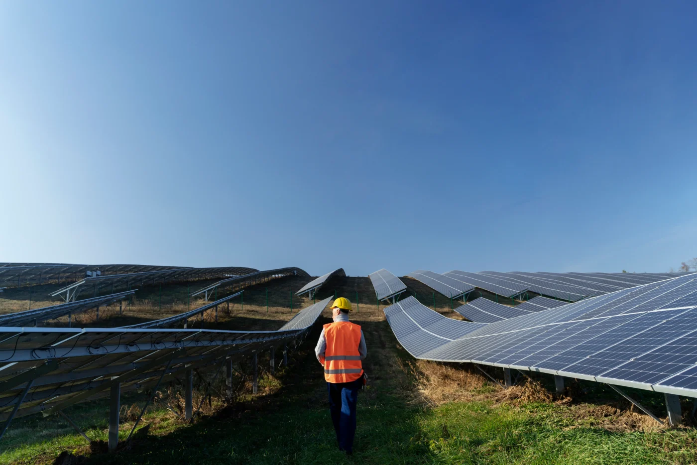 Técnico equipado com capacete e colete refletivo caminha entre fileiras de painéis solares, realizando inspeção visual em campo.