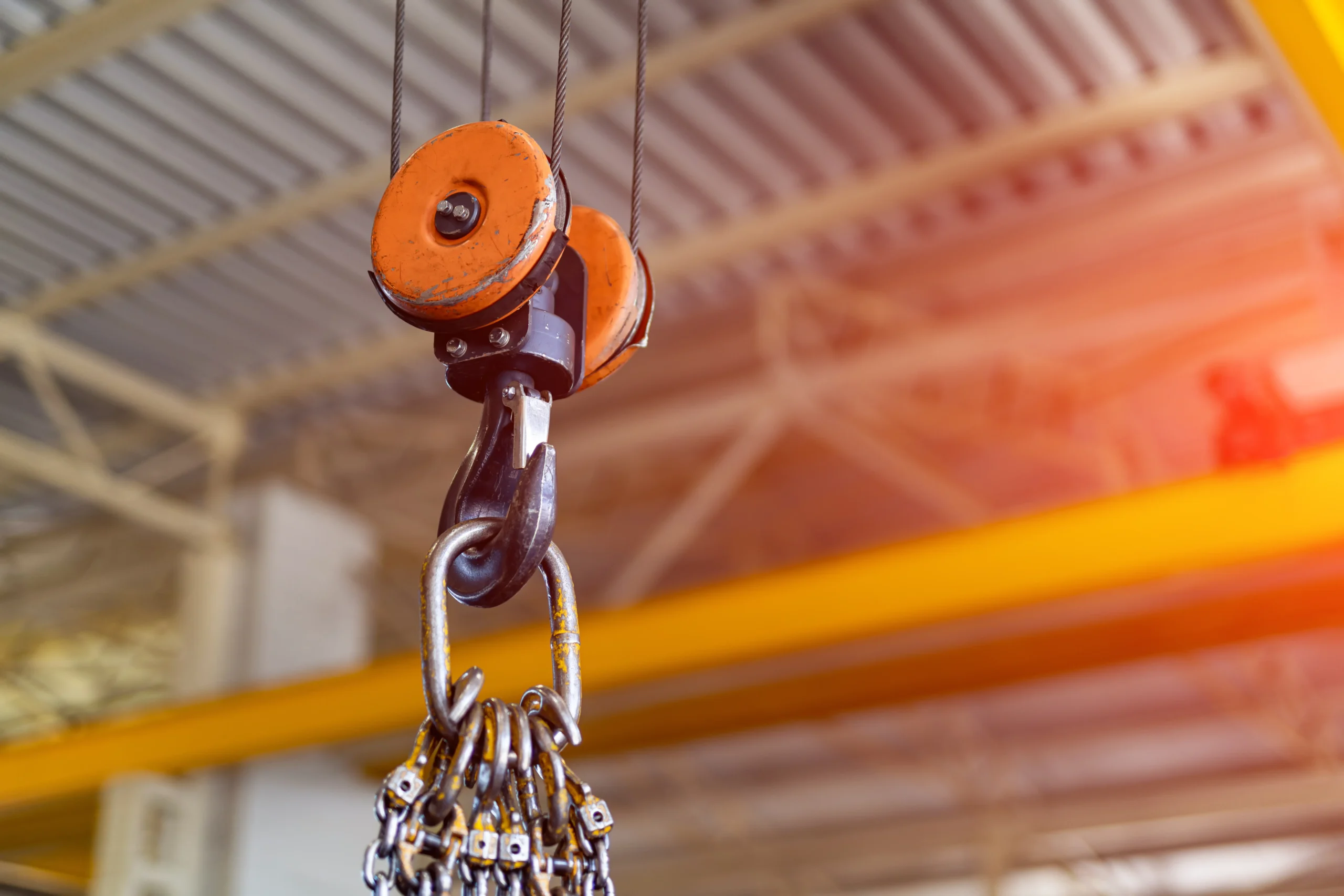 Imagem mostra uma talha com polia e gancho de elevação suspenso por cabos de aço em ambiente industrial, evidenciando componentes críticos do sistema de içamento, como roldanas, eixo, carcaça e ponto de fixação. A imagem remete à necessidade de inspeção periódica de desgaste, alinhamento, corrosão e integridade estrutural, fundamentais para a segurança operacional.