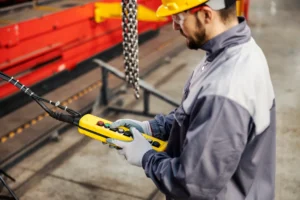 Profissional realiza inspeção funcional de talha elétrica por meio de comando pendente, verificando resposta dos controles, funcionamento dos botões de emergência e comportamento do sistema de elevação conforme critérios de segurança.