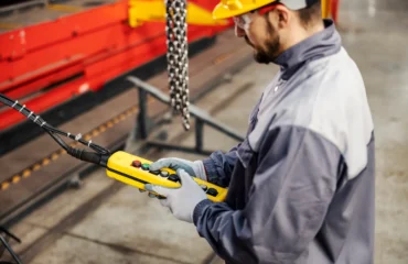 1 Profissional realiza inspeção funcional de talha elétrica por meio de comando pendente, verificando resposta dos controles, funcionamento dos botões de emergência e comportamento do sistema de elevação conforme critérios de segurança.