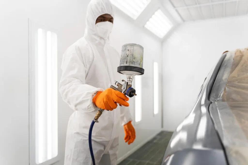 Aplicação de tinta em ambiente controlado com cabine pressurizada. Operador devidamente paramentado com EPI completo (macacão, máscara, luvas). Iluminação uniforme e paredes limpas indicam conformidade com boas práticas de pintura automotiva. - Laudo Cabine Pintura.