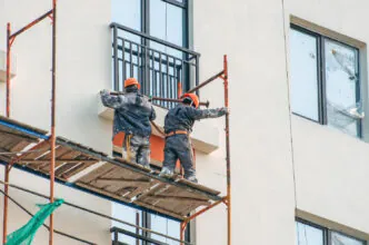 Trabalhadores realizam montagem de andaime em fachada de edifício, atuando em altura com uso de capacetes e vestimentas de proteção - Curso Acesso por Corda 2 NBR 15475
