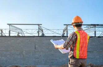 Um engenheiro ou supervisor de obra analisa plantas arquitetônicas em frente a um edifício em construção, assegurando que todos os detalhes estejam alinhados com o planejamento. - Curso Capacitação Incial NR 18. Curso NR 18 Inicial.