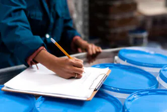 Registro de verificação de conformidade de embalagens durante processo de controle de qualidade em ambiente fabril. Profissional realiza anotação manual em checklist de inspeção.